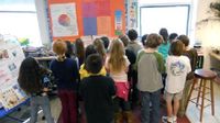A group of elementary students are huddling together in class, looking at a pie chart and bulletin board on the wall.