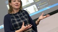 A closeup of a female teacher standing in front of a projector. It looks as if she's addressing her class, speaking, with her hands out, gesturing.