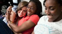 Three high school girls are sitting on a couch next to each other in class, smiling. The two girls to the right are hugging, and one of them is looking directly at the camera.