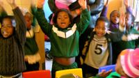 A group of eight young, elementary students are standing by their classroom desks with their arms raised in the air, talking. An adult is standing by them, holding a baby.