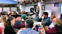 A group of adults are sitting among four tables, six to a table, in a library. Paper plates, popcorn bags, soda, and water bottles are on top of their tables, alongside open binders. A female adult is standing in front of everyone, addressing them.