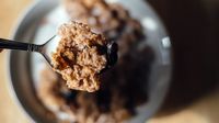 A closeup of a spoon with milk, corn flakes, and raisins. The rest of the image is out of focus. Beneath the spoon is a bowl filled with cereal on a light brown, wooden table.