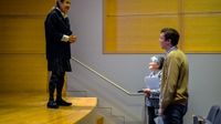 A woman is standing on a theater stage, looking down at a man and woman who are standing on the floor, close to the stage. They are all smiling at each other and talking.