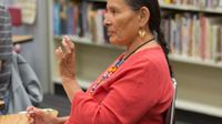 A side profile closeup of a female teacher sitting at a table in a classroom. A bookshelf behind her, and she's talking to someone. She's wearing a red shirt, a long, beaded necklace, and circular earrings.