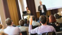 A group of teachers are sitting in a conference room, looking towards someone at a podium and someone standing in front of a big screen. One man in the audience has his hand raised.