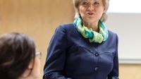 A short-haired blonde woman in her early sixties, wearing a blue coat and a blue, green, and yellow scarf, is standing in front of a projector, talking to a seated brunette woman wearing glasses.