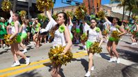 A street parade is going on in a downtown area. A lot of girls in white shirts and green shorts are walking in the street with their hands raised, carrying golden pom-poms.