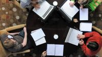 An aerial shot of five teachers sitting around a dark brown circular table in light brown cloth-covered chairs. There are three laptops, four notebooks, and three cups of coffee on the table.