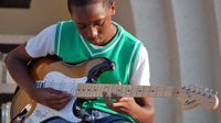 A young boy in jean shorts, a white t-shirt, and a green sweater vest is sitting outside in front of his school playing an electric guitar.