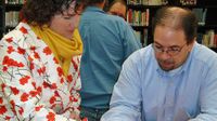 A woman in a floral sweater and a yellow scarf is standing, pointing to a piece of a paper on a table with her pen, and a man wearing glasses and a blue button-up shirt is sitting beside her, looking at the same paper. A bookshelf is behind them.