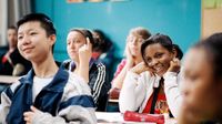 A group of high school students are sitting at desks in class, all looking towards the front, smiling.