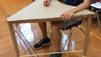 Chest down, a teen is sitting at a hand-made desk. It's made of a cardboard triangular desktop, three cardboard tubes as desk legs -- with tennis balls at the bottom of two of them -- and two solid, cylindrical wooden rods connecting the desk legs.