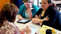 Three female teachers are sitting at a table, two on one side and one on the other, talking to each other and looking at their tablets.