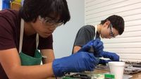 Two teenage boys in protective goggles, green aprons, and blue gloves are hovering over a desk holding a screwdriver, using it with some plastic-metal device.