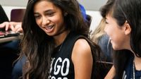 Two young, brunette, teenage girls are sitting in class, talking to each other and laughing.