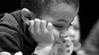 A black and white closeup of a young boy, maybe 5-years-old, in a hoodie. He's leaning forward with his head resting on his hand, and he has a solemn look on his face.
