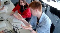 Teenage students are sitting at a long table in a classroom. One of them is talking into a microphone. They have a soundboard next to them with papers scattered across the table.