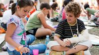 A group of young students are painting on paper, sitting outside on large pieces of cardboard laid out on the grass.