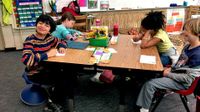 Four young students are sitting together at two, two-person desks put together as one table. They're near the front of the classroom near a whiteboard. The two girls are looking down, writing, and the two boys are looking up, smiling.