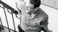A male teacher in glasses is leaning against a stairway rail, smiling.