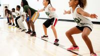 Seven young students are squatting side-by-side on a basketball court with their arms extended out at their sides.