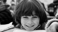 A closeup of a young kid's face, smiling, with their chin perched on their desk next to an open textbook.
