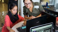 A female adult wearing a green shirt and glasses is sitting beside a young female student in a red blouse. They're both looking at a laptop computer.