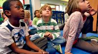 A group of young students are sitting on the floor in their classroom, looking up.