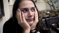A closeup of a teenage girl sitting slouched, resting her head in her cupped hands. She's looking up and to the side with tears in her eyes. She's sitting in a naturally lit room with couches, comfy chairs, coffee tables, and a small, potted tree.
