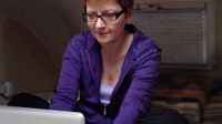 A red-headed woman in a purple sweatshirt with glasses is hunched over, sitting cross-legged, typing on her laptop.