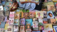 A young black girl is laying down on top of about 300 books stacked together on top of each other, looking up at the camera, smiling.