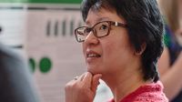 A side profile, closeup of a female teacher with her hand in a fist against her chin. She's in a pink sweater and wearing brown glasses.