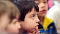 A closeup of a young boy cupping his face with his hands, sitting among other students on the floor.