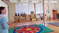 A female teacher is standing at the left side of a classroom. The classroom is empty, except for a large green and brown rug placed side by side with two benches placed on top of them, and shelves are against the back wall with drums on top of them.