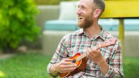 Bearded man sitting outside, smiling, playing the ukulele