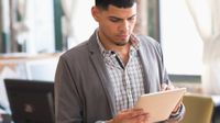 A young man in a checkered button up shirt and a gray sweater is standing in a classroom, holding and looking down at a silver tablet.