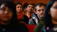 High school students are sitting in red chairs in a theatre. A teenage boy, two rows back from the front, is in focus; everyone else is slightly blurry.