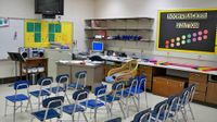 17 chairs are lined side-by-side in three rows facing the front of a classroom. At the front of the class, inches from the ceiling, a flat screen TV is attached to the wall. A countertop with drawers extends along the right side of the room.