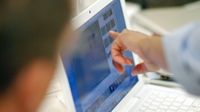 Someone's forearm is in the shot, and their index finger is touching a laptop screen, pointing at a frame within a video editing software. Another person's head is in the shot, blurred, looking at the computer screen.