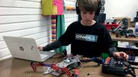 A young boy wearing a black, long-sleeve shirt and headphones has one arm extended next to his laptop, and the other hand is touching a remote control. In between his laptop and the remote control is a robotic device made out of plastic, metal, and wires.