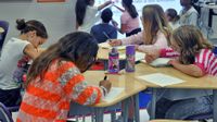 Four young girls are sitting, writing at their desks. Five young students are standing by a projector at the front of the class, pointing at the screen, which is projecting four different sentences marked with blue marker.