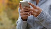 A closeup of a young woman's hands holding an iPhone. She's wearing a grey, full zip sweatshirt, and the autumn-colored background is blurred out.