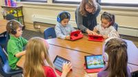 Five young elementary students are sitting around a six-sided table in a classroom. They're all wearing headphones and playing on iPads. A female teacher is assisting one of them.