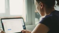 A closeup of a young woman, facing away from us, sitting down next to a window, typing on her laptop. Light is shining into the room from the window.