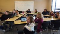 Ten students are sitting in a classroom at rectangular tables positioned in a triangle, all looking up in the same direction.