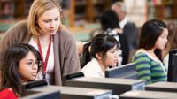 A librarian is peering over the shoulder of a student who is sitting next to two other female students, all working on desktop computers in a library.