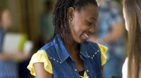 A teenage girl in a yellow shirt and jean vest is smiling, looking down at an opened binder in her hands, showing it to another girl outside of the frame of the photo. The rest of the photo is blurred out, and seems to be students in a school hallway.