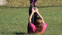 A teenage female in a pink t-shirt, black exercise pants, and purple tennis shoes is lying on the grass with her legs raised straight up and her hands reaching towards her feet.