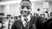 A young boy in a white collared shirt, tie, and sweater is smiling directly into the camera.