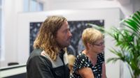 A side profile of a male and female adult from the waist up standing in a spacious white-walled room looking in the same direction. There's a black and white painting behind them, and a green, potted plant beside them.